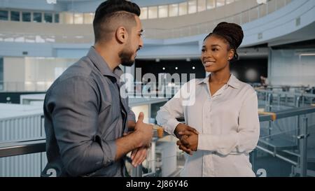 Friendly multiethnic business partners colleagues african woman and indian arabian man talking standing in modern company office hall two diverse Stock Photo