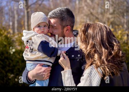 A family with small baby. Father holding daughter on his hands. Baby watching at the camera. Mother holding child's hand. Stock Photo