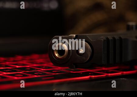 The gun lies on a black laptop keyboard with red backlight, security threat, cyber attack, system hacking Stock Photo