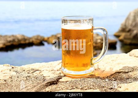 Glass of light beer near sea, close-up. Misted mug, pint of cold beer stands against background of blurred sea landscape in cafe, restaurant, bar, pub Stock Photo