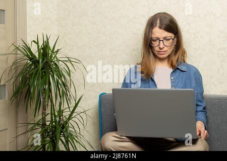 A young woman works on a laptop while sitting on the couch at home. Stock Photo
