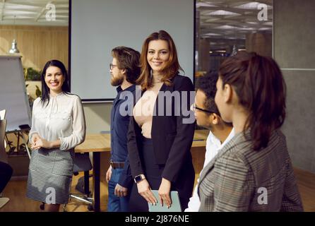 Beautiful smiling young business woman standing surrounded by colleagues in meeting at office. Stock Photo