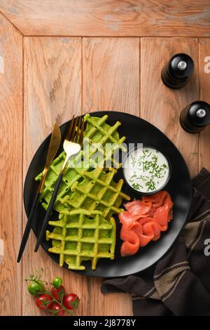 Green Belgian waffles. Spinach or wild garlic or pesto waffles with red salmon and cream sauce on black plate over dark rustic wooden background. Deli Stock Photo