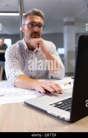 Caucasian male engineer using laptop while sitting with blueprint at desk in modern workplace Stock Photo