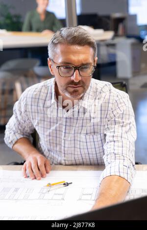 Confident caucasian male engineer using laptop while sitting with blueprint at desk in workplace Stock Photo