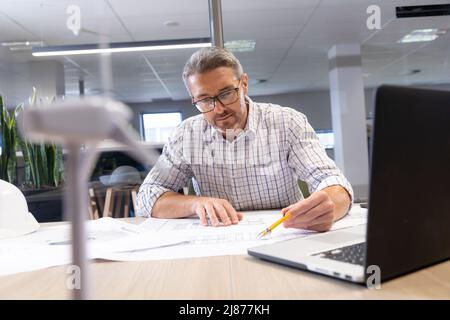 Confident caucasian male engineer analyzing blueprint at desk in modern workplace Stock Photo
