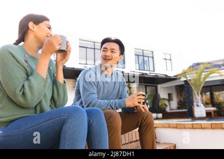 Happy young asian couple chatting while having coffee outside house, copy space Stock Photo
