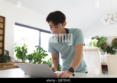 Asian teenage boy with headphones using laptop on table while standing at home, copy space