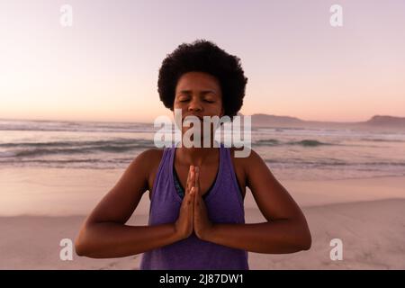 African american mature woman with afro hair meditating in prayer pose against sea and clear sky Stock Photo