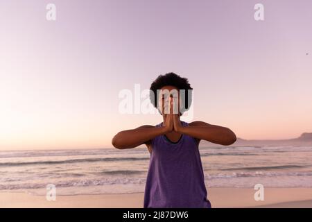 African american mature woman with afro hair meditating in prayer pose at beach against clear sky Stock Photo