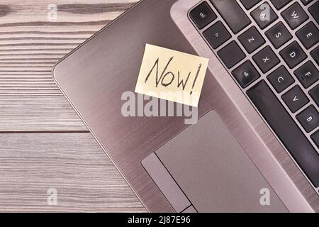 Sticky note with now handwritten now word on the laptop pc. Top view computer on grey wood. Stock Photo
