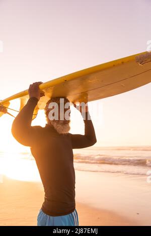 African american senior man carrying surfboard on head while standing against sea and clear sky Stock Photo