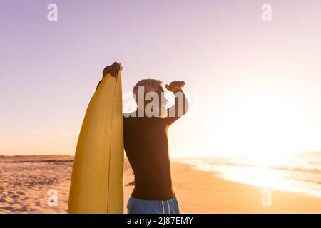 African american bearded senior man with surfboard shielding eyes while looking at sea at sunset Stock Photo