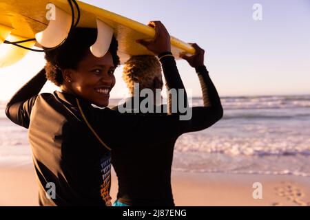 Portrait of smiling african american mature woman with man carrying surfboard on head at beach Stock Photo