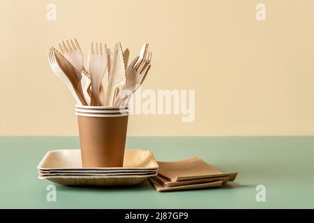 Eco-friendly disposable tableware against green beige background. Wooden forks and knives in paper cups on bamboo plates. Single use tableware. Stock Photo