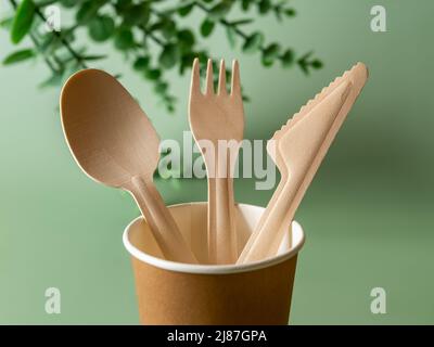 Eco-friendly wooden fork, knife and spoon in a brown paper cup against green background. Biodegradable dishes for picnics, fast food, takeaways. Stock Photo