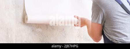 A man rolls out a roll of white wallpaper on the wall with glue. Wallpapering. Repair of a room, apartment, house. Stock Photo