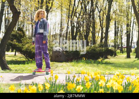 cute Caucasian blonde curly-haired young girl standing on a skateboard in the park full shot outdoors . High quality photo Stock Photo