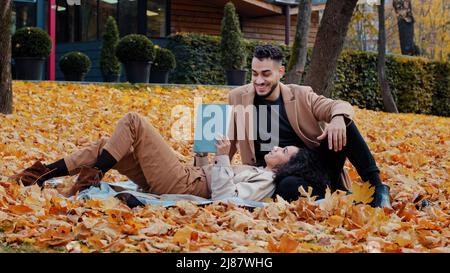 Date outdoors young bearded arab guy sincerely talking with beloved woman man nods approvingly beautiful girl lying on plaid in yellow leaves reading Stock Photo