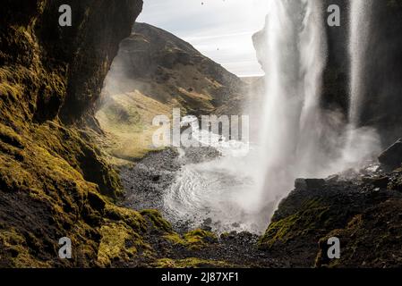 Beautiful view from behind the Kvernufoss waterfall through its misty cascade and spray, bathed in magical light, near Route 1 / Ring Road, Iceland Stock Photo