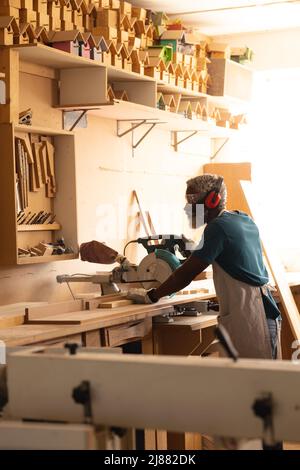 African american mature carpenter in earmuffs and protective eyewear using circular saw in workshop Stock Photo