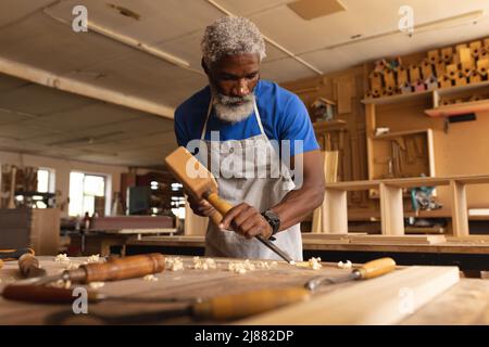 African american mature carpenter using chisel and mallet on plank at workbench in workshop Stock Photo