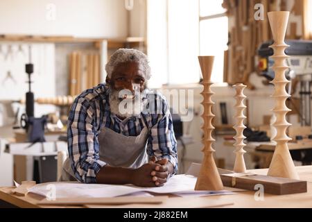 Portrait of smiling african american mature carpenter leaning on workbench at workshop Stock Photo