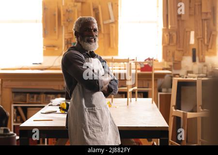 Portrait of smiling african american mature carpenter standing with arms crossed in workshop Stock Photo