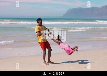 Cheerful african american young man holding hands and spinning daughter at beach against sky Stock Photo