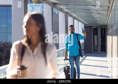 Happy african american young businessman taking selfie through smartphone with luggage at corridor Stock Photo