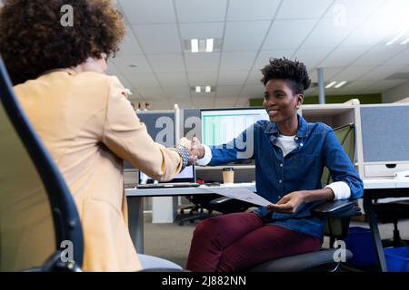Smiling african american businesswoman shaking hands with hispanic female colleague in cubicle Stock Photo