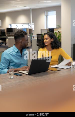 Biracial young female and male colleagues discussing together while sitting at desk in workplace Stock Photo
