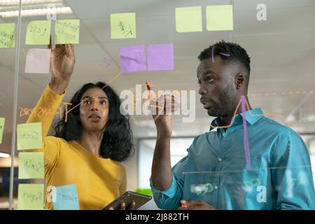 Confident biracial male and female colleagues discussing over strategy on glass wall at workplace Stock Photo