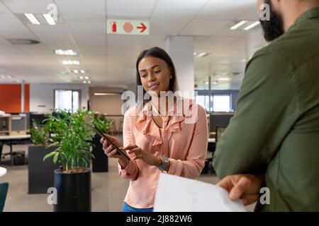 Smiling biracial businesswoman discussing over tablet pc with caucasian businessman at workplace Stock Photo