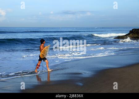 A Caucasian man carrying a yellow long board surfboard and leaving the sea with waves in the background at Batu Bolong Beach, Canggu, Bali. Stock Photo