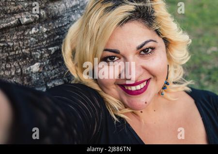 close up portrait beautiful blonde plus size caucasian young woman smiling taking a selfie outdoors sitting on grass, looking at camera, lifestyle con Stock Photo