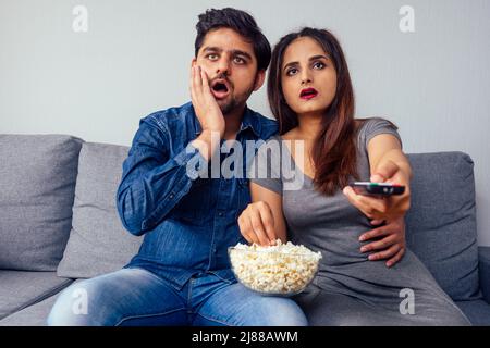 amazed young indian couple watching thriller movie with remote control in hand in living room with popcorn Stock Photo