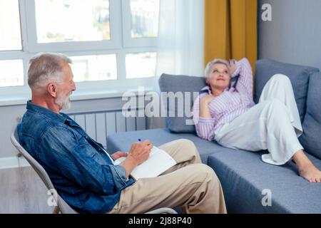 beautiful short haired goog-looking woman lying on the couch,old man artist sitting next to her on chair and drawing in day light living room with big Stock Photo