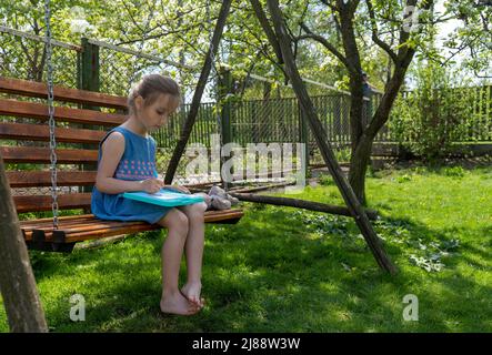 Adorable little girl is concentrated on learning to write while sitting on a swing on a sunny day Stock Photo