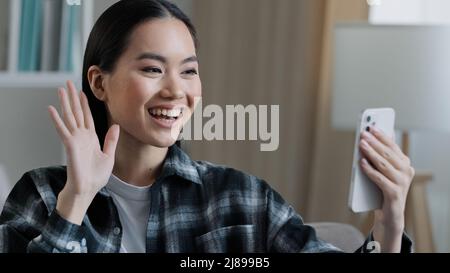 Happy friendly asian girl blogger vlogger smiling female distant woman mobile phone video call remote communication greeting waving hand talking Stock Photo