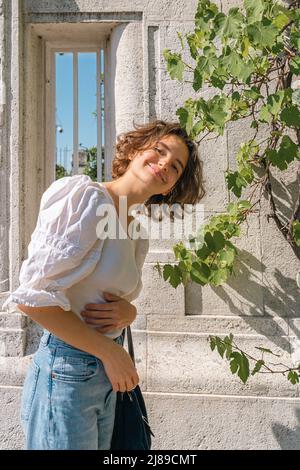 Young happy teenage girl with brown curly hair smiling while looking at camera, against backdrop of ancient European-style architecture. Positive smil Stock Photo