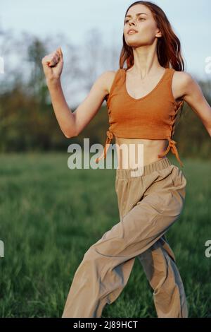 A woman works out and runs through the field in the park with a smile in a good mood looking at the beautiful summer nature around her Stock Photo
