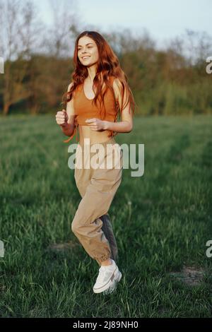 A woman works out and runs through the field in the park with a smile in a good mood looking at the beautiful summer nature around her Stock Photo