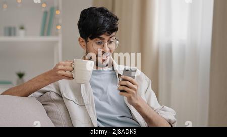 Millennial arabic guy indian handsome 30s man sitting at home on couch drinking hot tea coffee cappuccino cocoa from cup receive message looking at Stock Photo