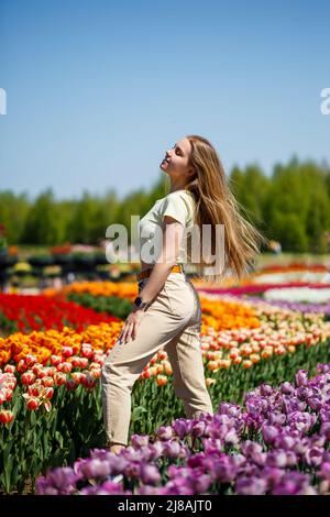 Beautiful woman in hat laughing to camera in spring flower garden. Girl with camera smiling in park with many flowers. Woman enjoying time in blooming Stock Photo