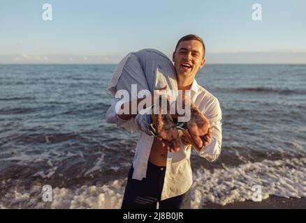 Portrait of laughing man holding woman on his shoulder, standing on beach by sea Stock Photo