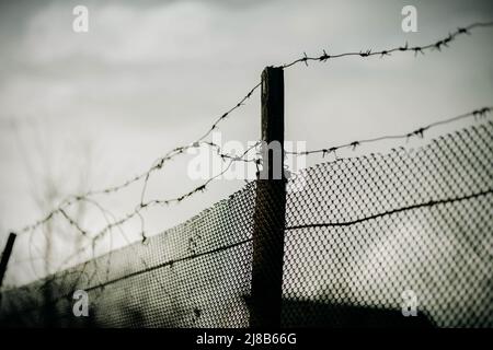 There is an old fence with a terrible barbed wire against the background of a sad gray overcast sky. War and prison. Stock Photo