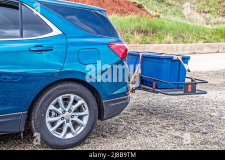 Blue hatchback car parked on gravel with hitch cargo carrier holding tied down rubber storage box on back - Closeup and cropped Stock Photo