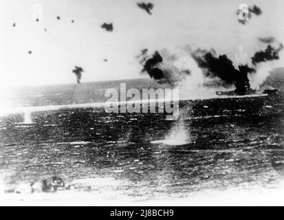 The U.S. Navy aircraft carrier USS Lexington (CV-2) under air attack on 8 May 1942 during the Battle of the Coral Sea, as photographed from a Japanese plane. Heavy black smoke from her stack and white smoke from her bow indicate that the view was taken just after those areas were hit by bombs. The destroyer in the lower left appears to be USS Phelps (DD-360). Stock Photo