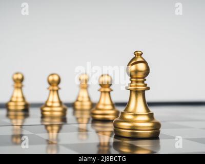 The golden bishop chess piece in front of gold pawns on chess board, white background. Bishop represents the church, important and powerful entity thr Stock Photo